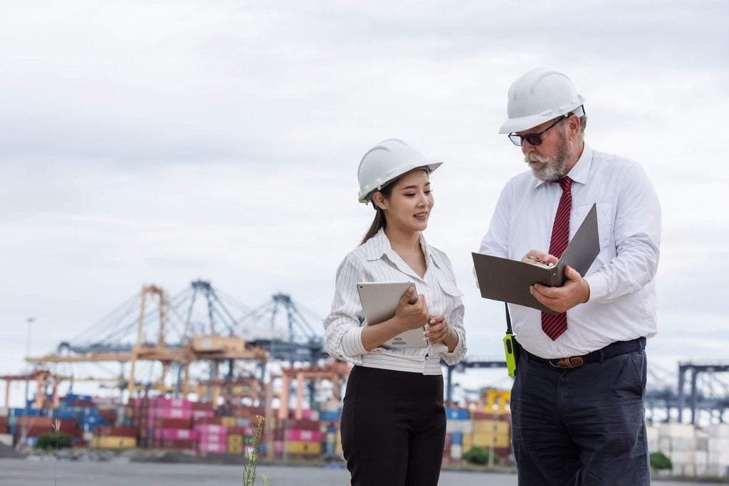Two people in safety helmets and business attire discuss documents at a shipping port, with colorful cargo containers and cranes visible in the background, as they review progress on Multi-Agent DA Reconciliation processes.