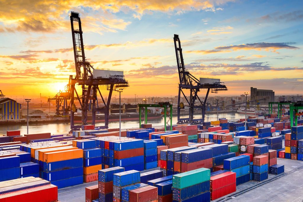 Shipping containers stacked at a busy port with large cranes and a colorful sunset sky in the background, illustrating the efficiency of port agency quotation services in managing complex logistics.