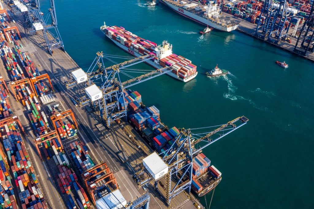 Aerial view of a busy shipping port with large cranes, stacked colorful shipping containers, cargo ships, and tugboats in the water—a scene where accurate port agency quotes ensure smooth operations.