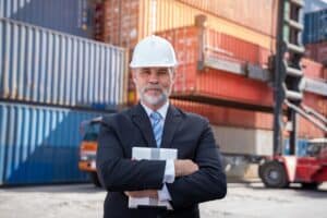 A man in a suit and white hard hat stands with arms crossed, holding a tablet, in front of stacked shipping containers at an industrial shipping yard.
