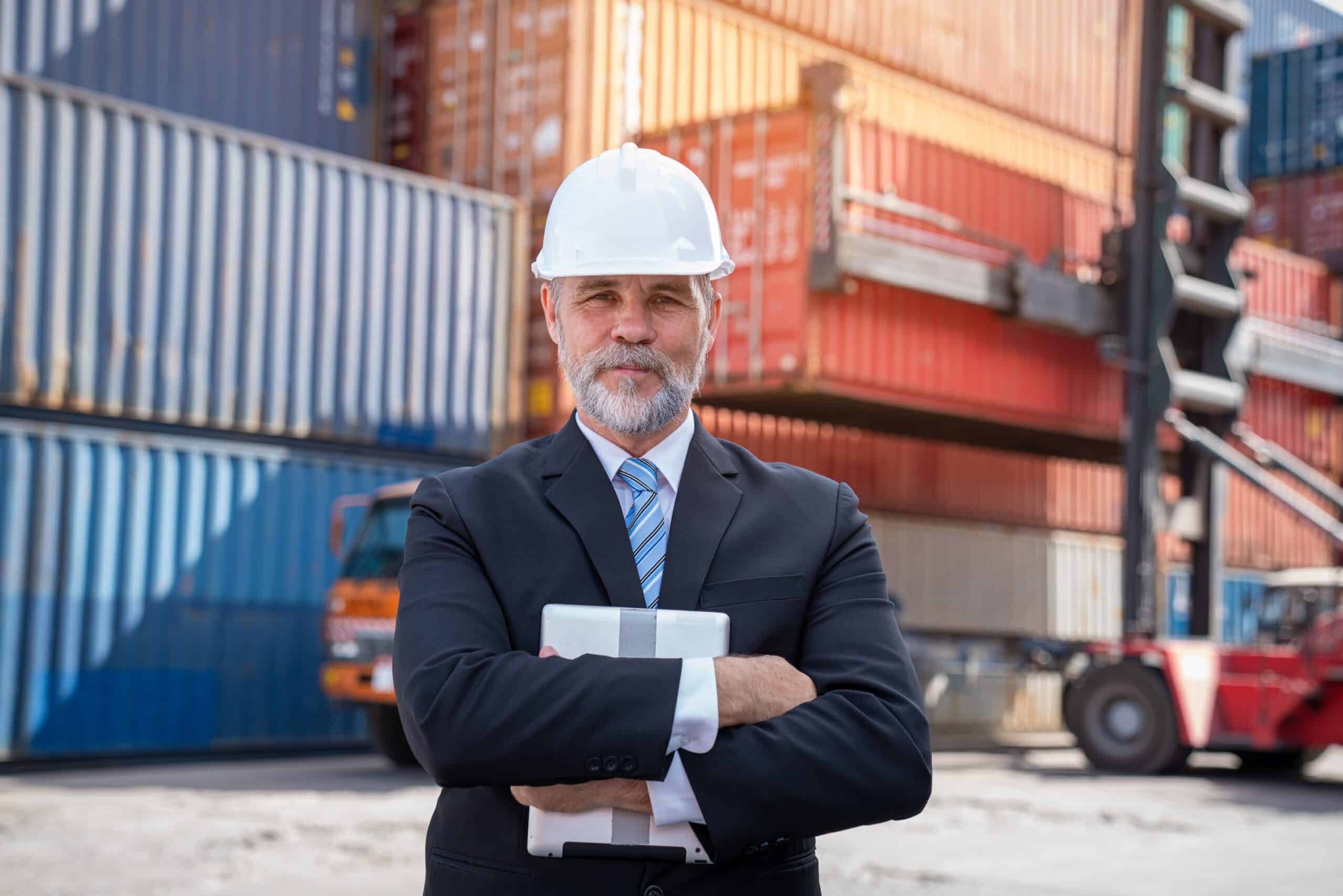 A man in a suit and white hard hat stands with arms crossed, holding a tablet, in front of stacked shipping containers at an industrial shipping yard.