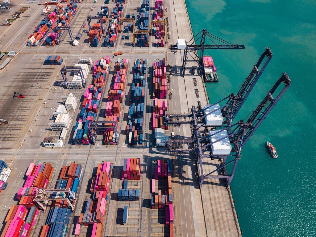 Aerial view of a shipping terminal where wharfage charges apply, featuring stacked cargo containers, industrial cranes, and a small boat in the turquoise water.