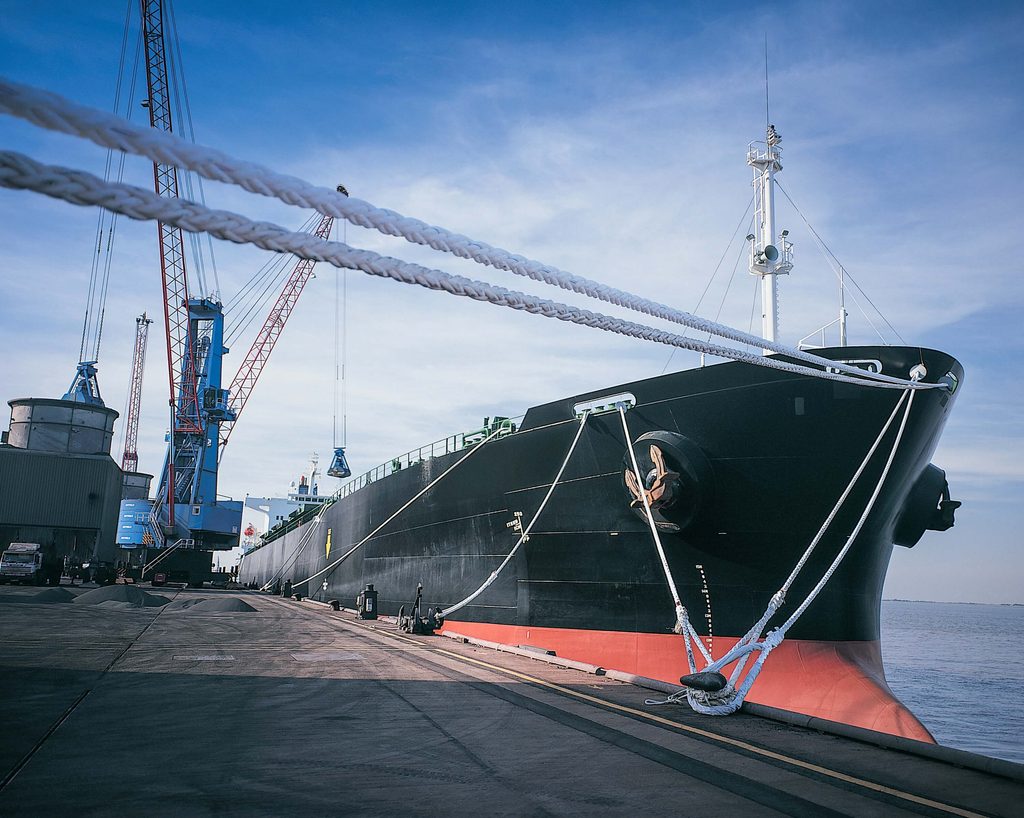 A large black and red cargo ship moored at a dock, secured by thick ropes, with industrial cranes visible in the background.