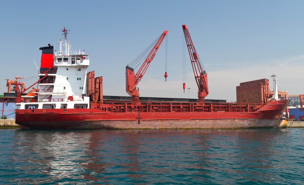 A red and white cargo ship with two cranes docked in a harbor.