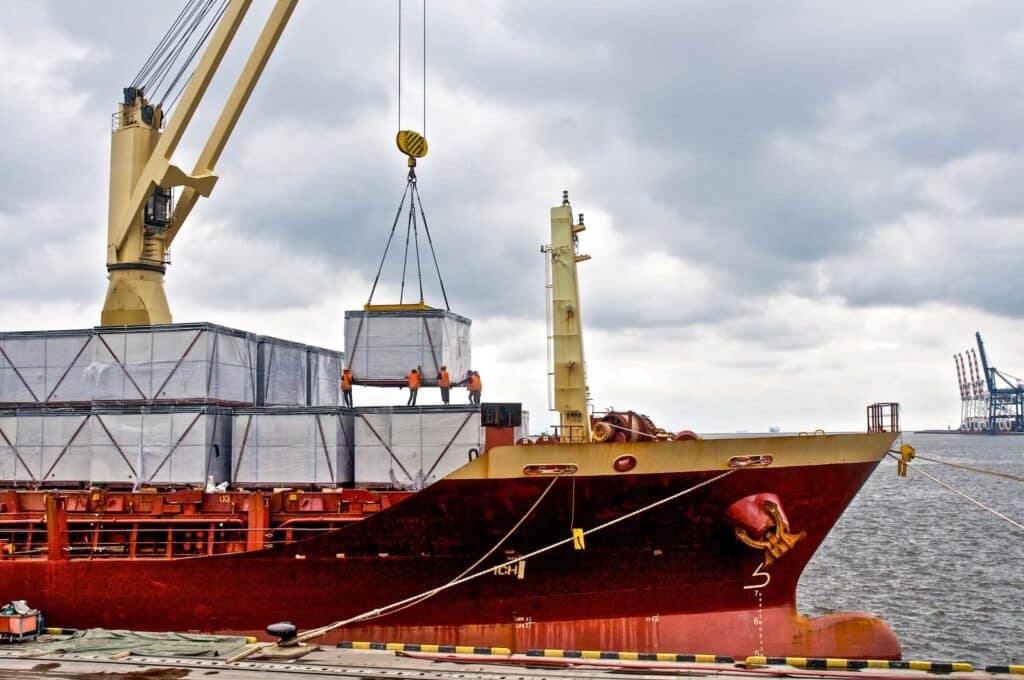 A crane lowers a large, wrapped cargo container onto the deck of a red cargo ship while workers in high-visibility vests guide it into place.