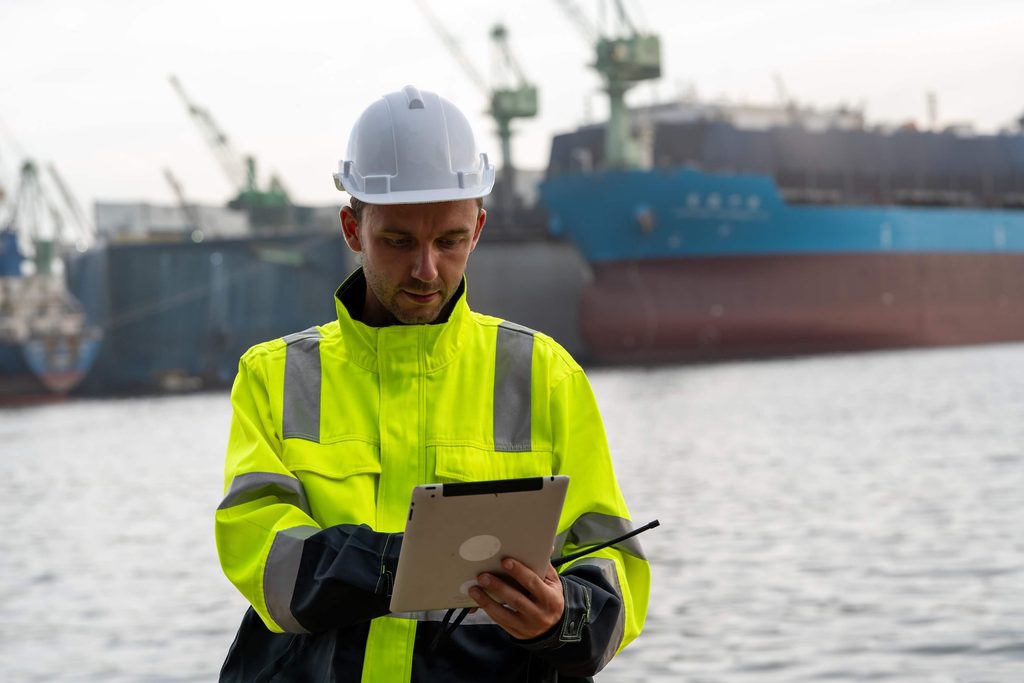 A worker in a white hard hat and a high-visibility yellow jacket uses a tablet in front of a shipyard with large vessels.