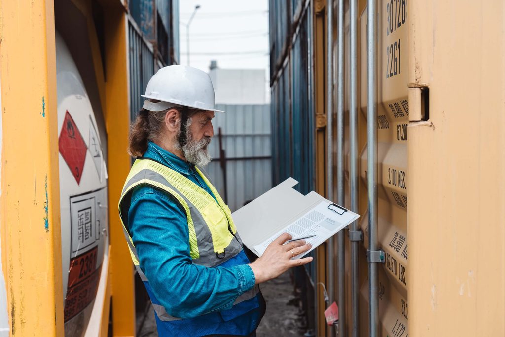 A worker in a hard hat and high-visibility vest inspects a shipping container and documents wharfage charges on a clipboard.
