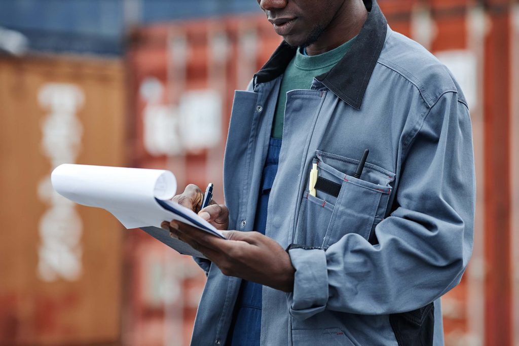 A worker in a gray uniform writes on a notepad near shipping containers while calculating wharfage charges.